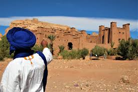 Man in a blue turban pointing toward a desert mud-brick fortress